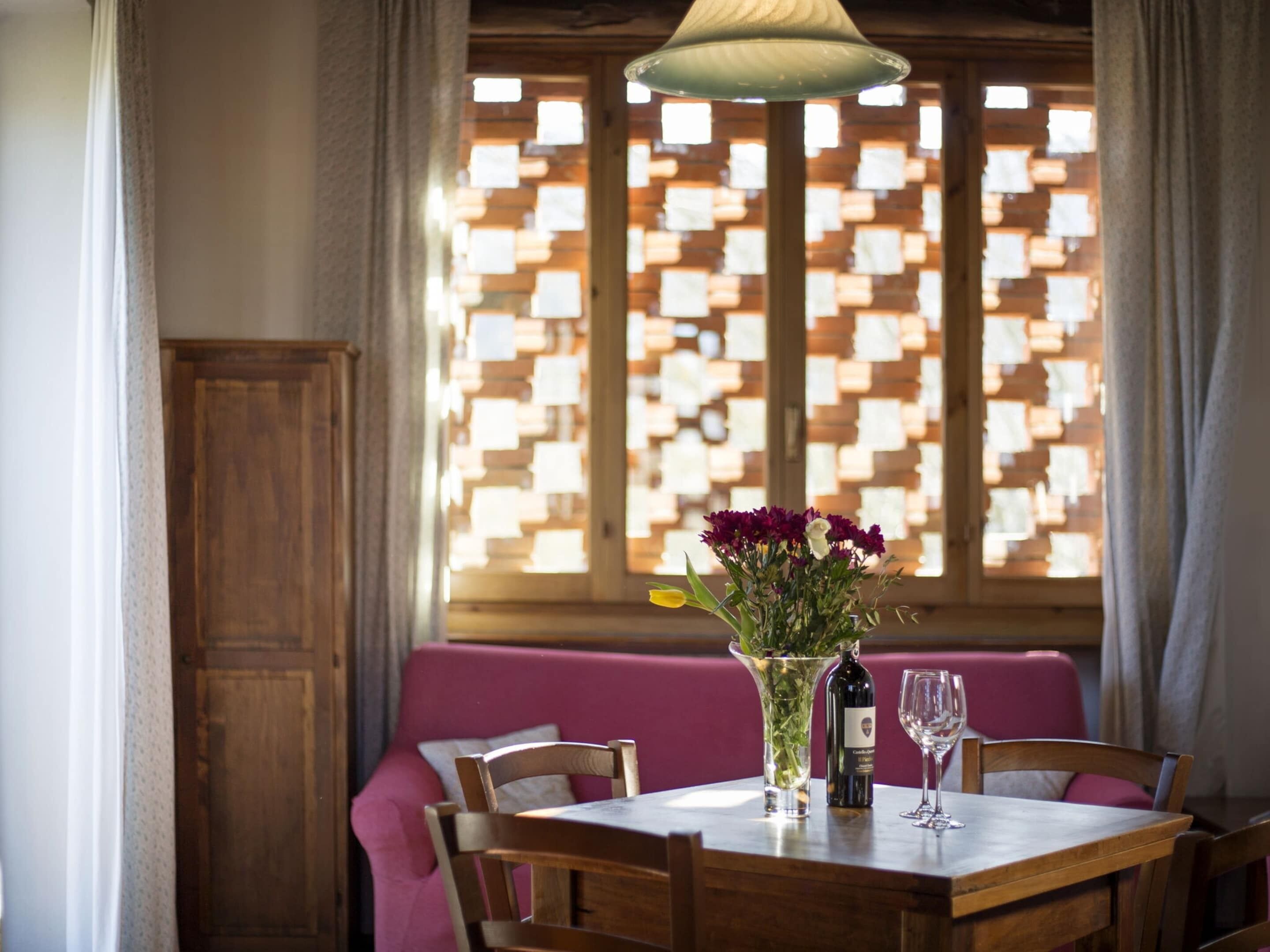 A dining area with a table set for two, a vase of flowers, a bottle of wine, and a glass. The room has wooden walls and a window with a patterned blind.