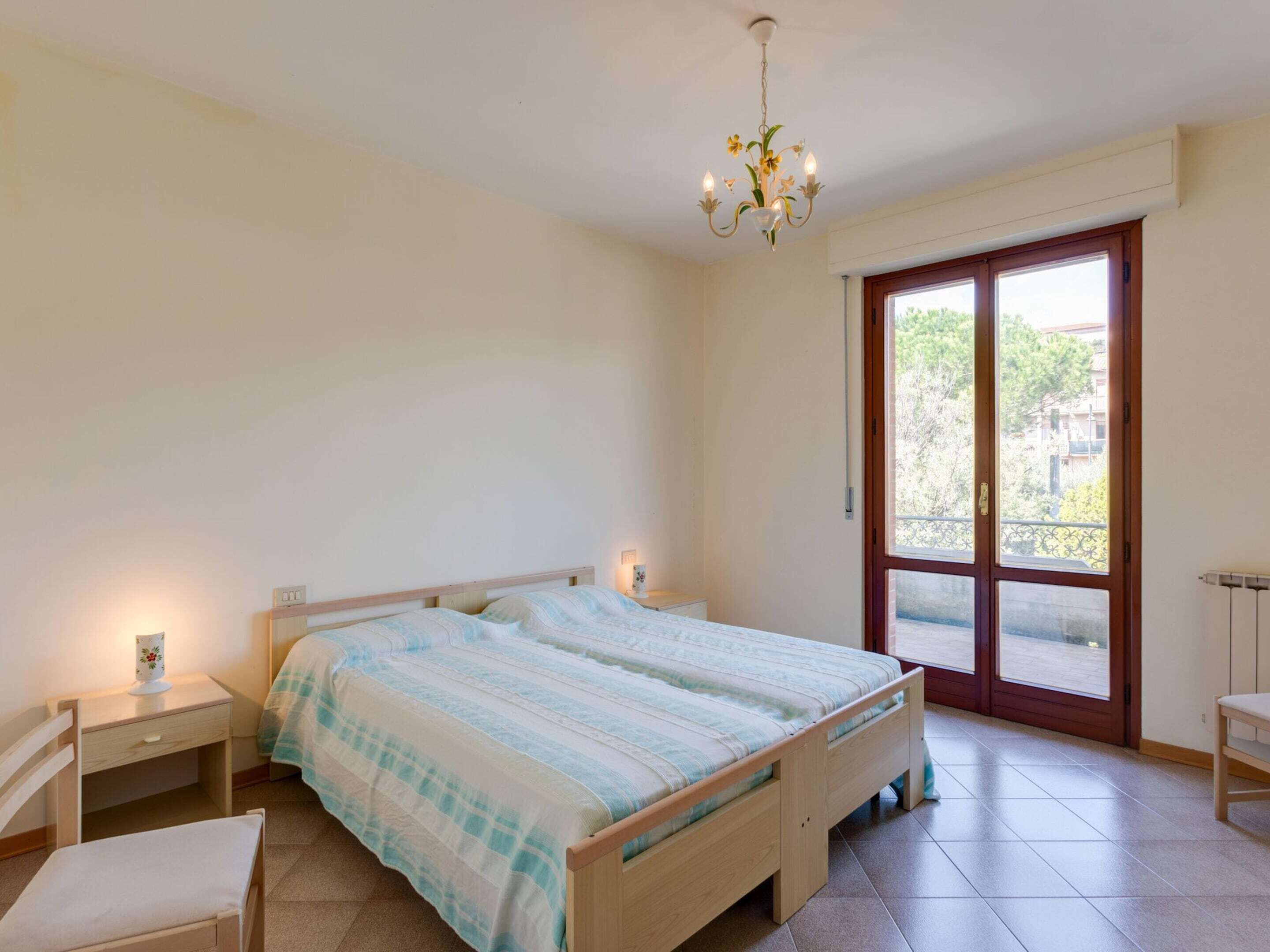 A bedroom with a double bed, a nightstand, a chair, a window with a view, and a chandelier.