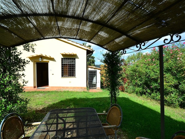 Un patio cubierto con una mesa y sillas, un edificio blanco con una puerta y una ventana, y un jardín con follaje y flores.