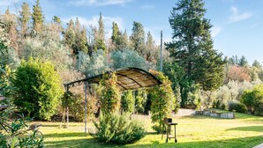 A covered outdoor area with a pergola, a small table, and a cart, surrounded by lush greenery and trees. - Belvilla by OYO Loggia (San Casciano in Val di Pesa)