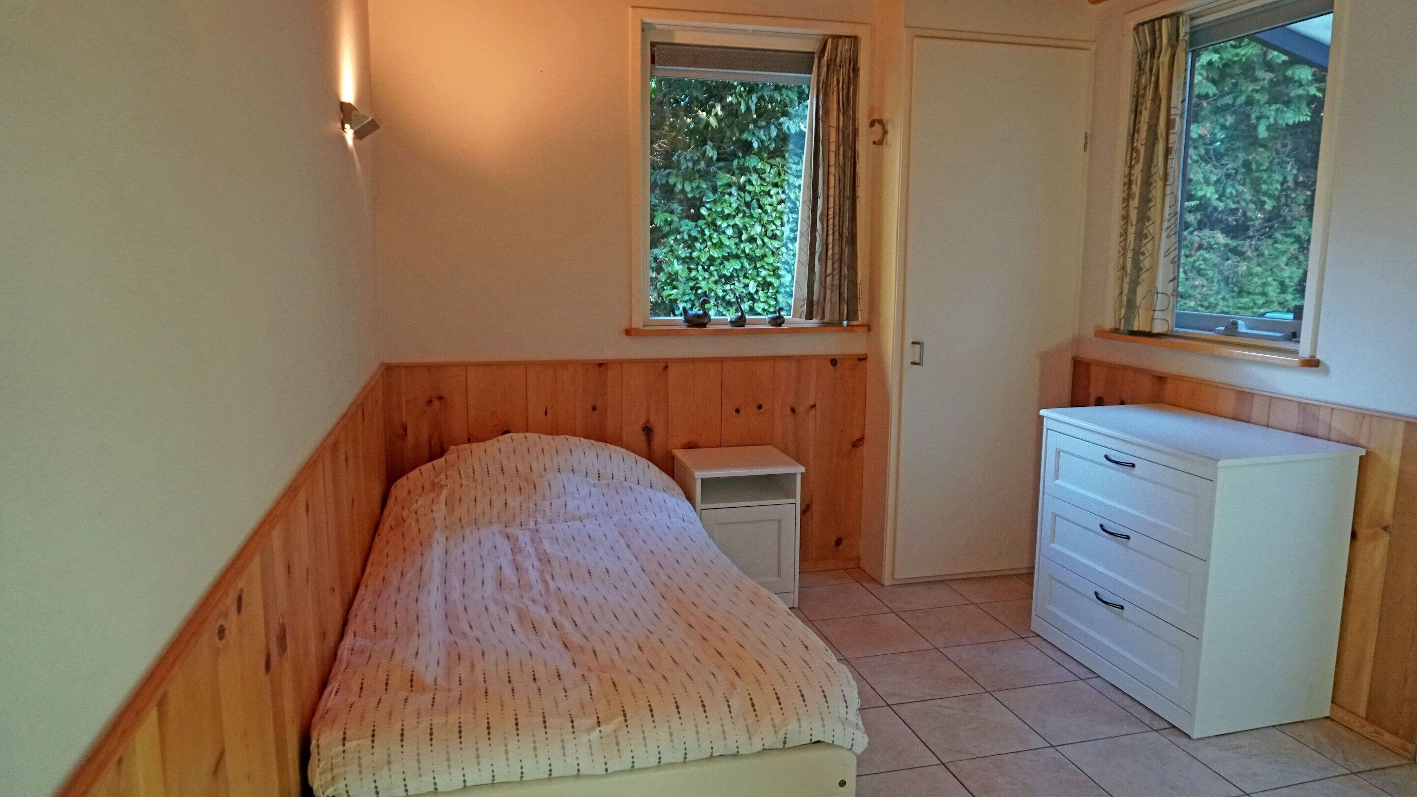 Een slaapkamer met een bed, nachtkastje, dressoir en een raam met uitzicht op groen.