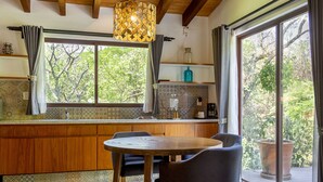 A kitchen with wooden cabinets, a round table, and chairs, a window with curtains, and a view of greenery outside. - Hotel Amate del Rio (Malinalco)