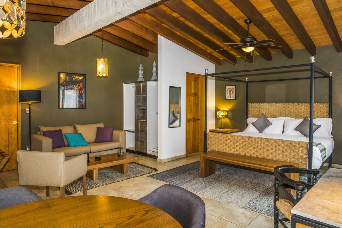 A modern bedroom with a four-poster bed, a sofa, a dining table, and a ceiling fan.