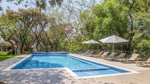A rectangular swimming pool with a wooden deck, surrounded by lounge chairs and umbrellas, set in a lush garden with trees. - Hotel Amate del Rio (Malinalco)