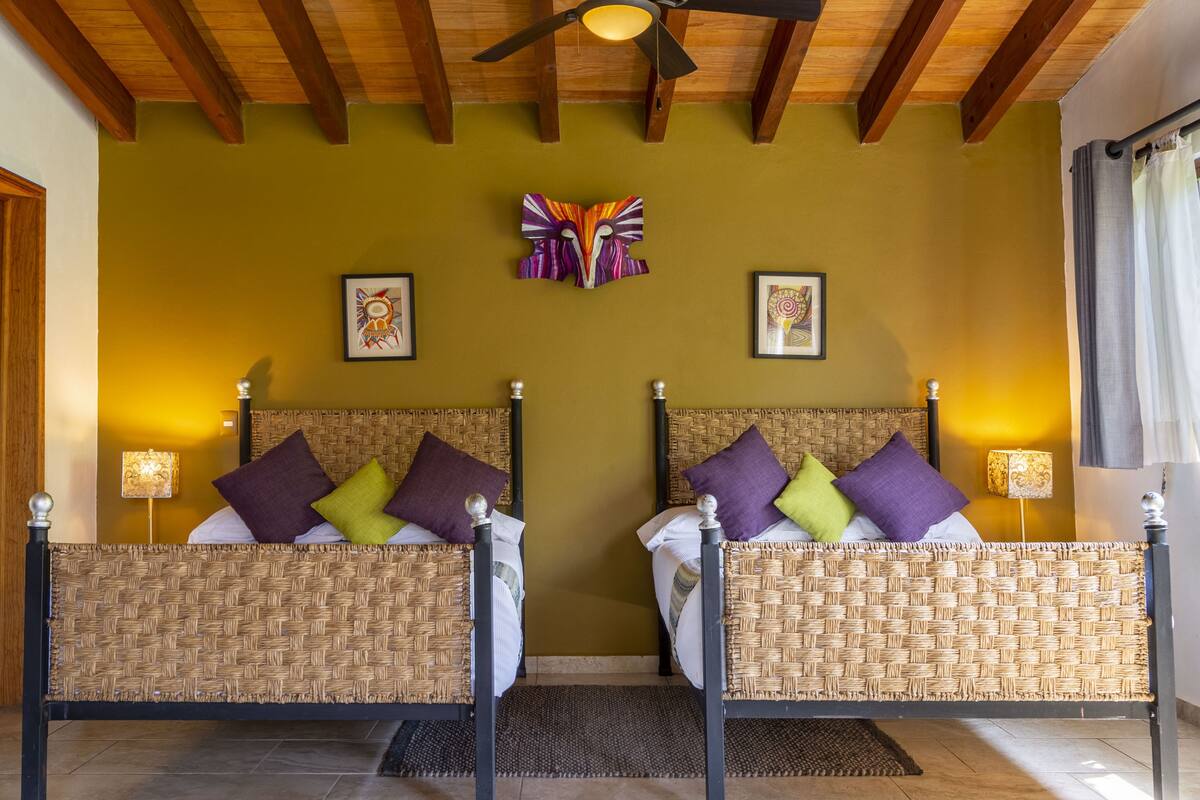 A room with two beds, each with a woven headboard, a ceiling fan, and wall-mounted artwork.