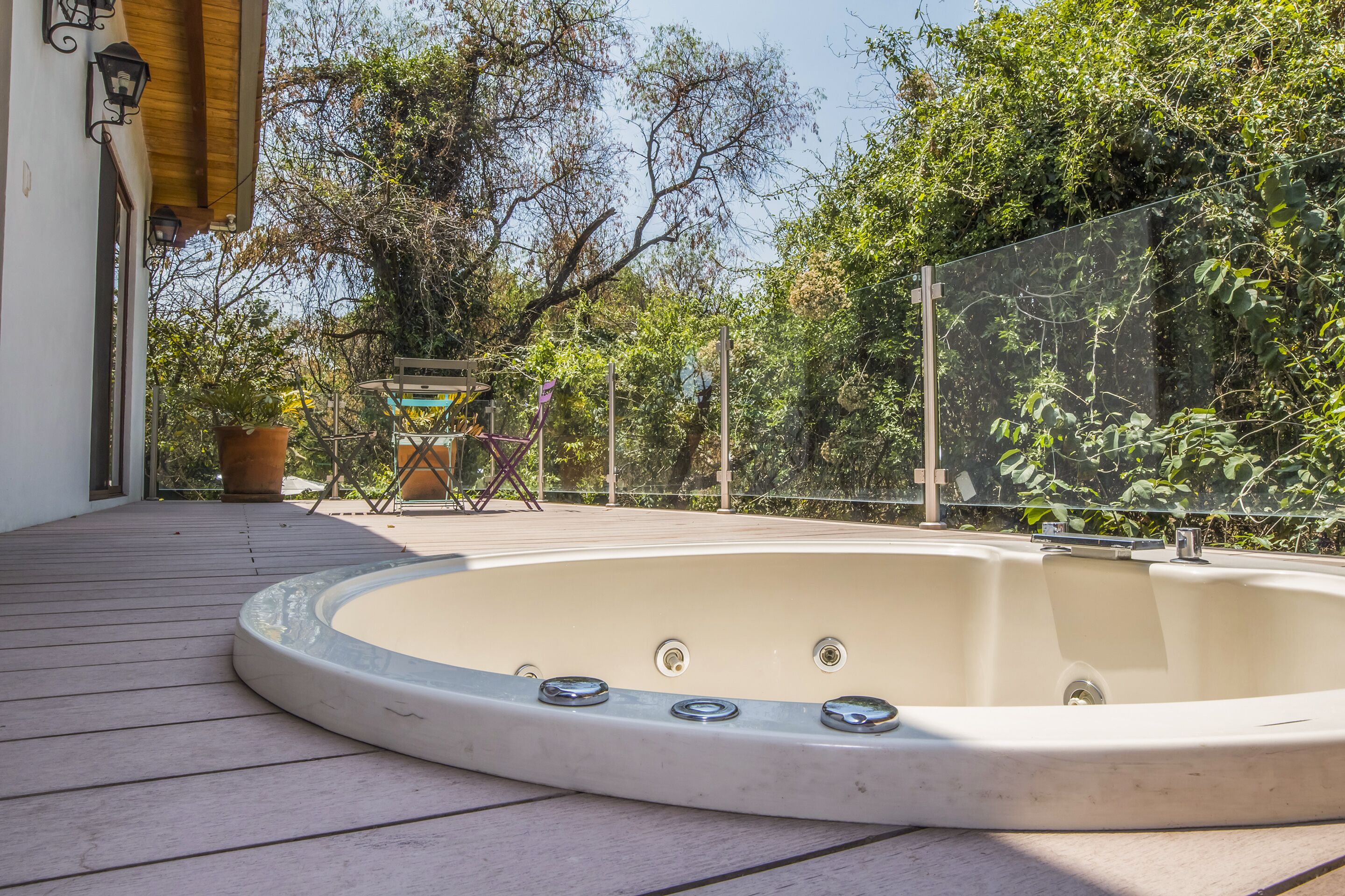 A hot tub on a wooden deck with a glass railing, surrounded by greenery and a building with a white facade.