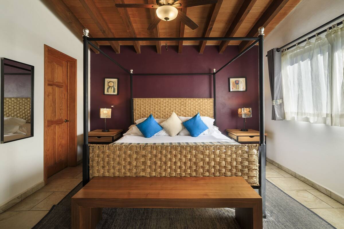 A four-poster bed with a woven headboard, wooden floor, and a ceiling fan.