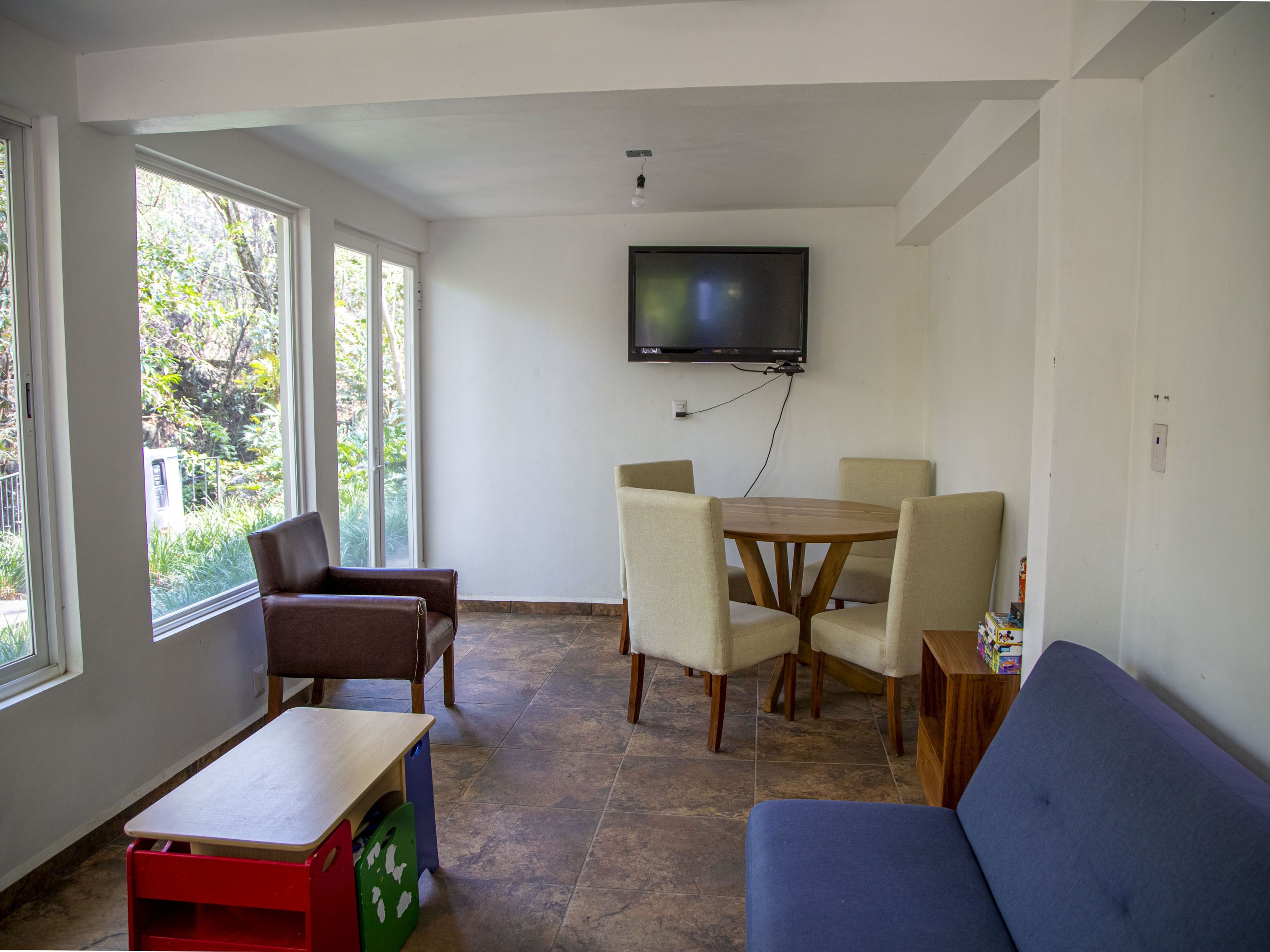 A room with a TV mounted on the wall, a dining table with chairs, a sofa, and a small table with a bench.