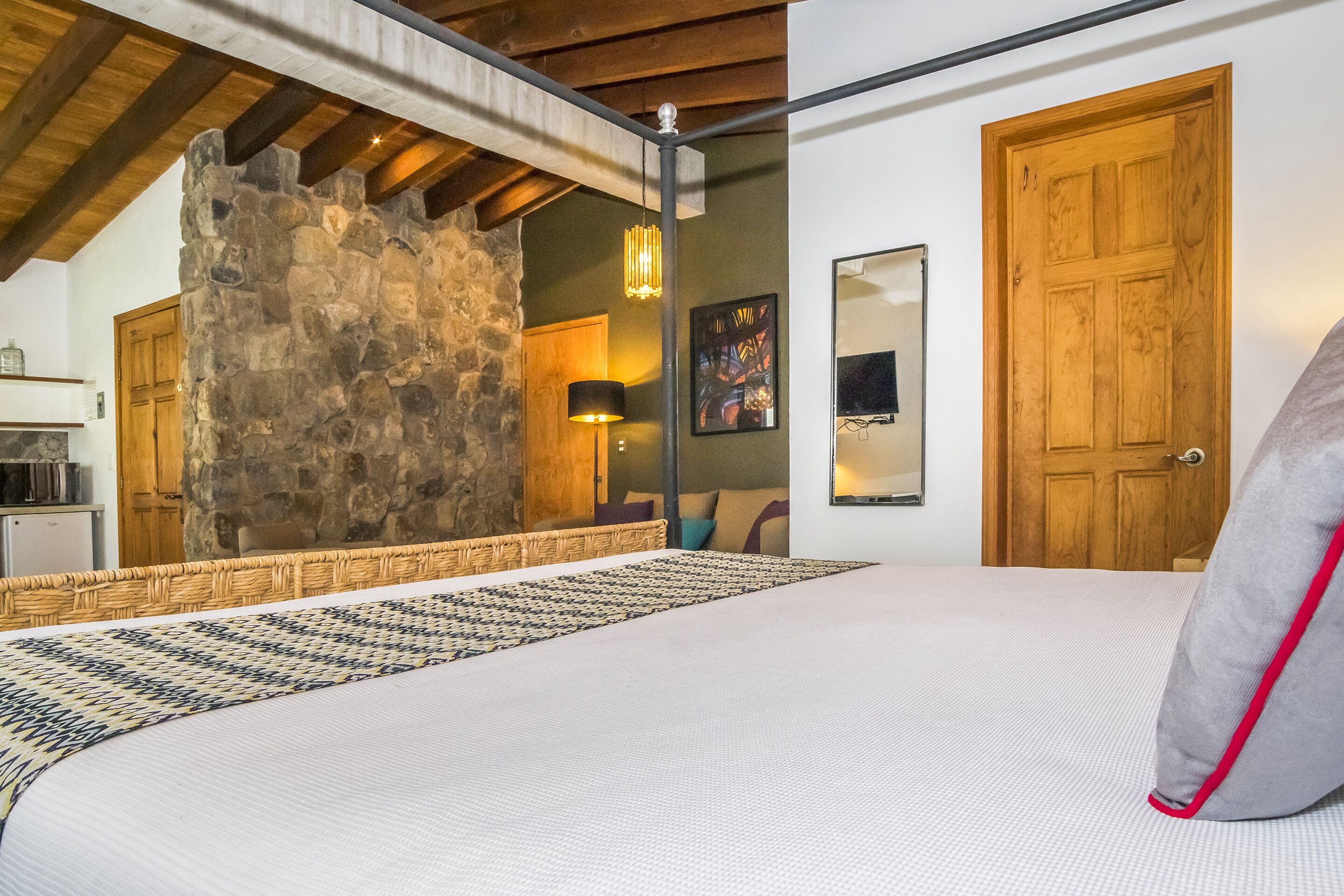 A bedroom with a stone wall, wooden ceiling beams, a bed with a patterned bedspread, a bedside lamp, and a wooden door.