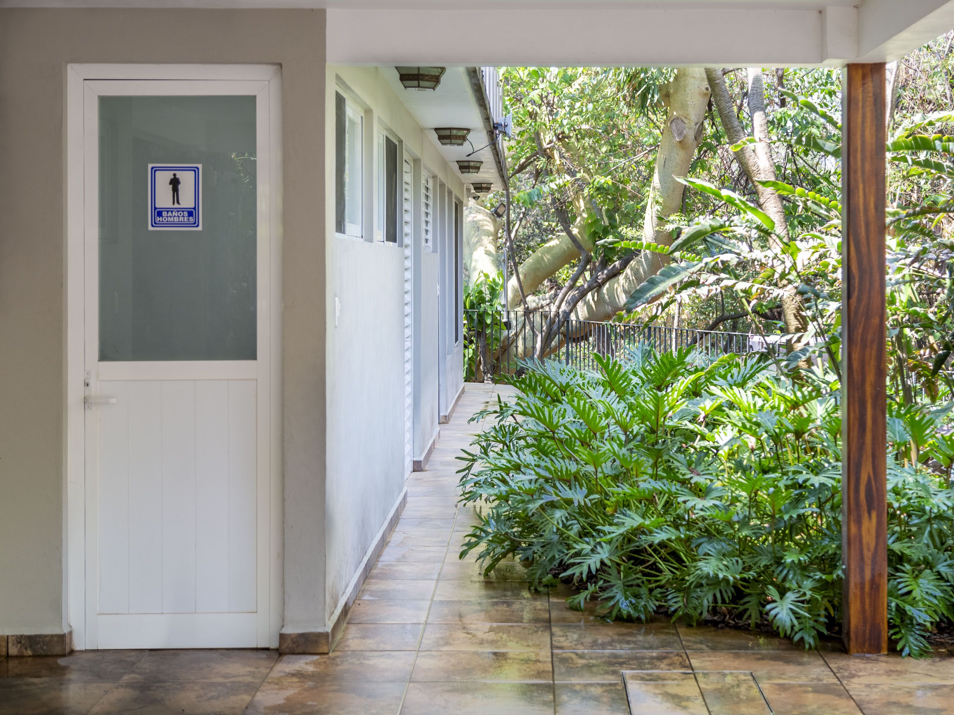 A corridor with a door on the left, a sign indicating a men's restroom, and a tiled floor leading to a lush green garden area.