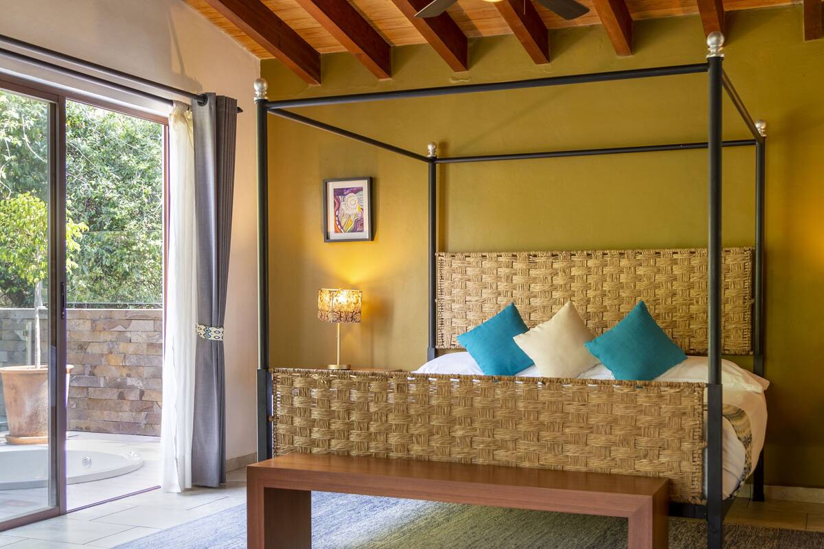 A four-poster bed with a woven headboard, a bedside table with a lamp, and a bench in a room with a view through a glass door.