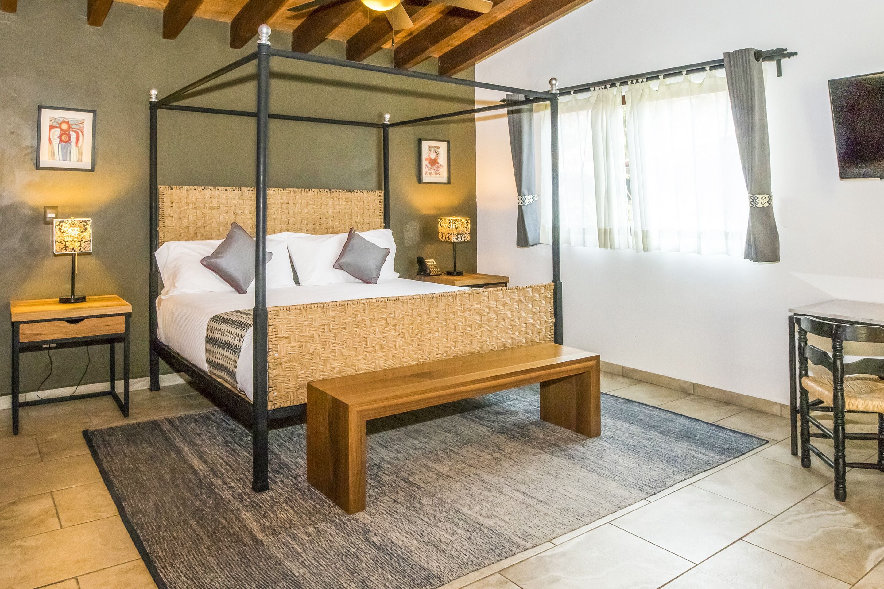 Uma cama de dossel com cabeceira em tear, mesas de cabeceira com abajures, um banquinho e vista para o exterior através de uma janela com cortinas.