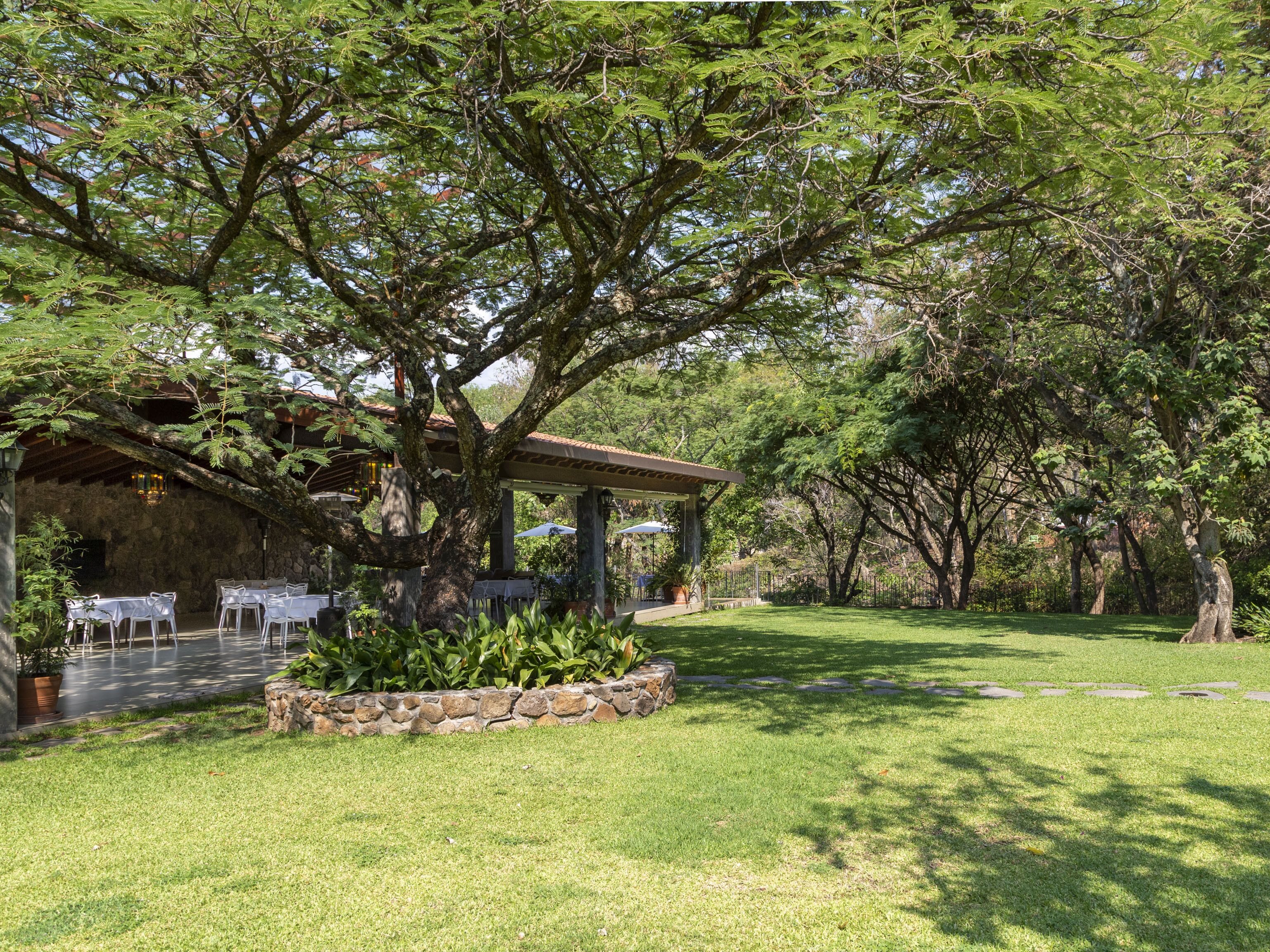 A covered outdoor seating area with stone walls and tables, surrounded by lush greenery and trees.