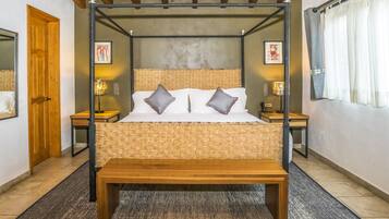 A four-poster bed with a woven headboard, two bedside tables with lamps, a wooden bench, and a door to the left.