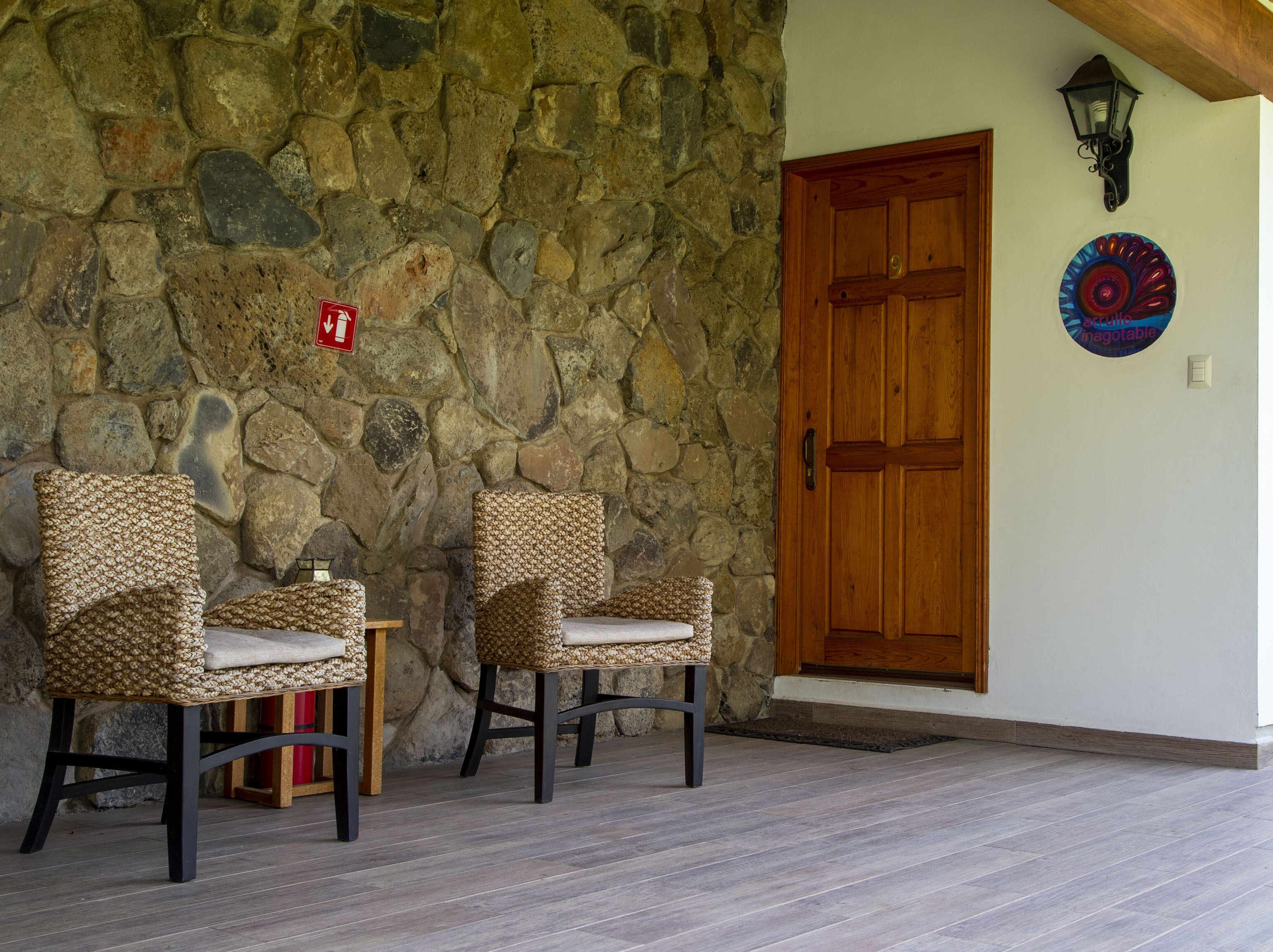 Un mur de pierre, deux fauteuils en osier, une porte en bois et un luminaire fixé au mur.