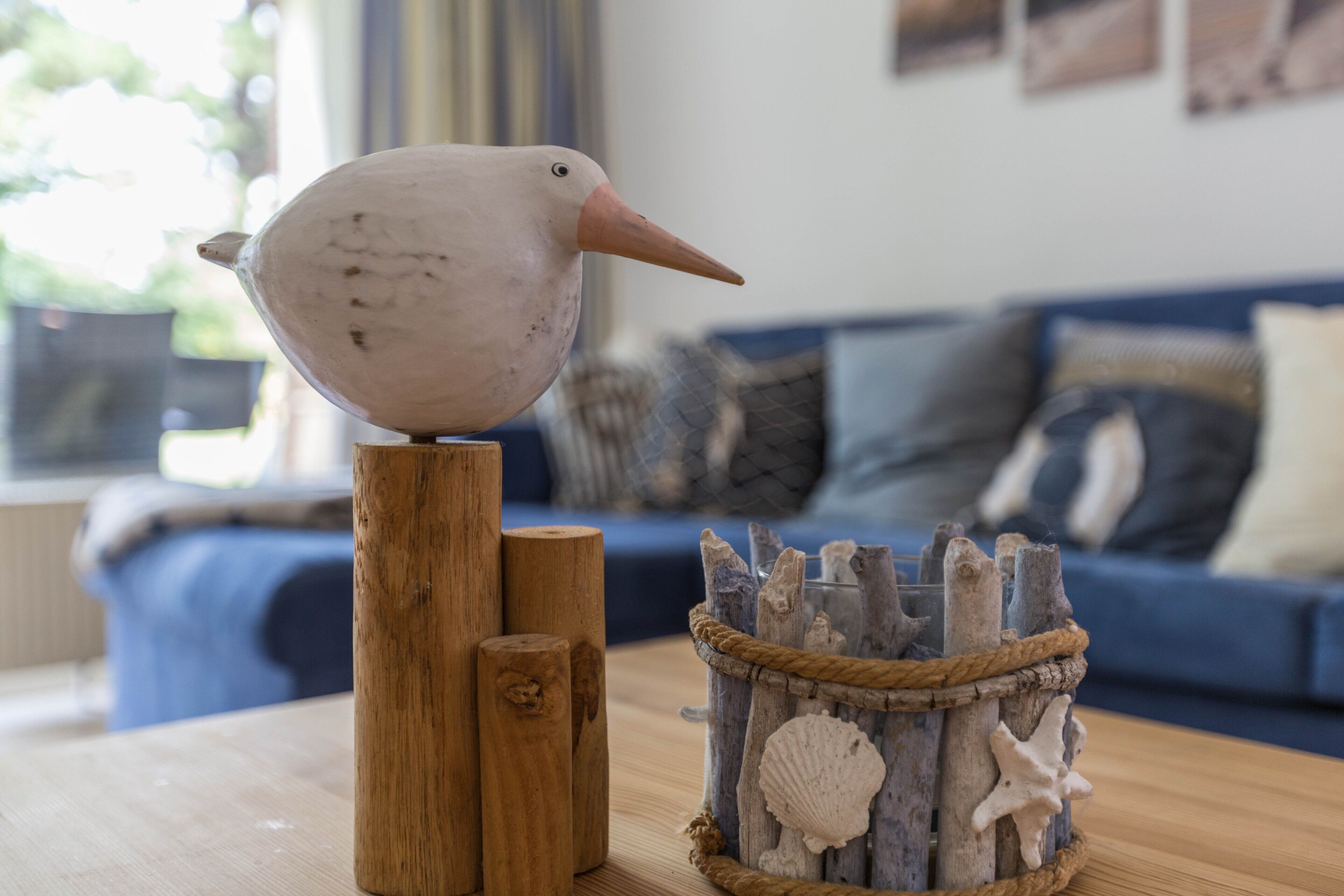 Eine Vogel-Figur auf einem Holzständer, ein rustikaler Korb mit Muscheln und eine blaue Couch im Hintergrund.