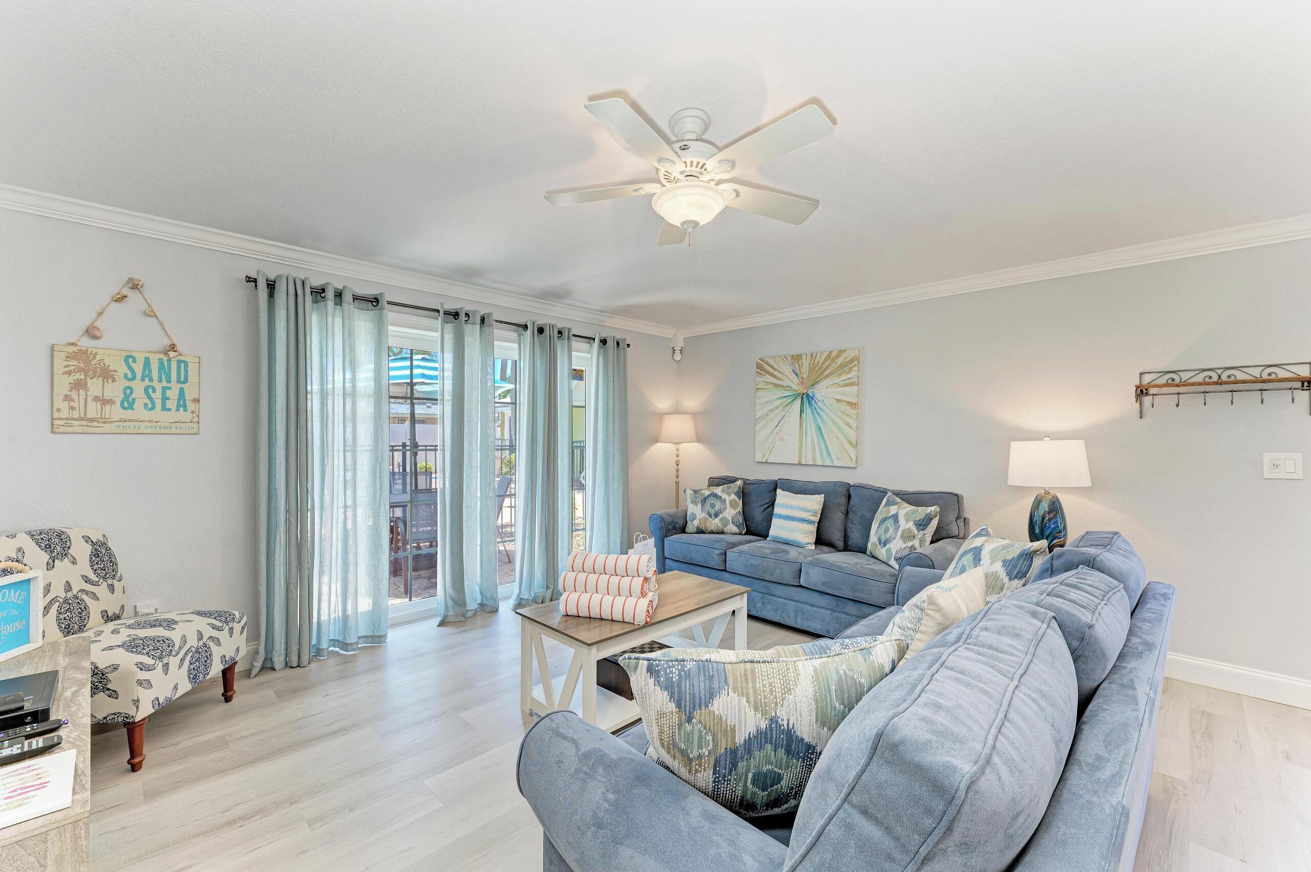 A living room with a blue sofa, a coffee table, and a ceiling fan. There is a sign on the wall that reads "SAND & SEA."