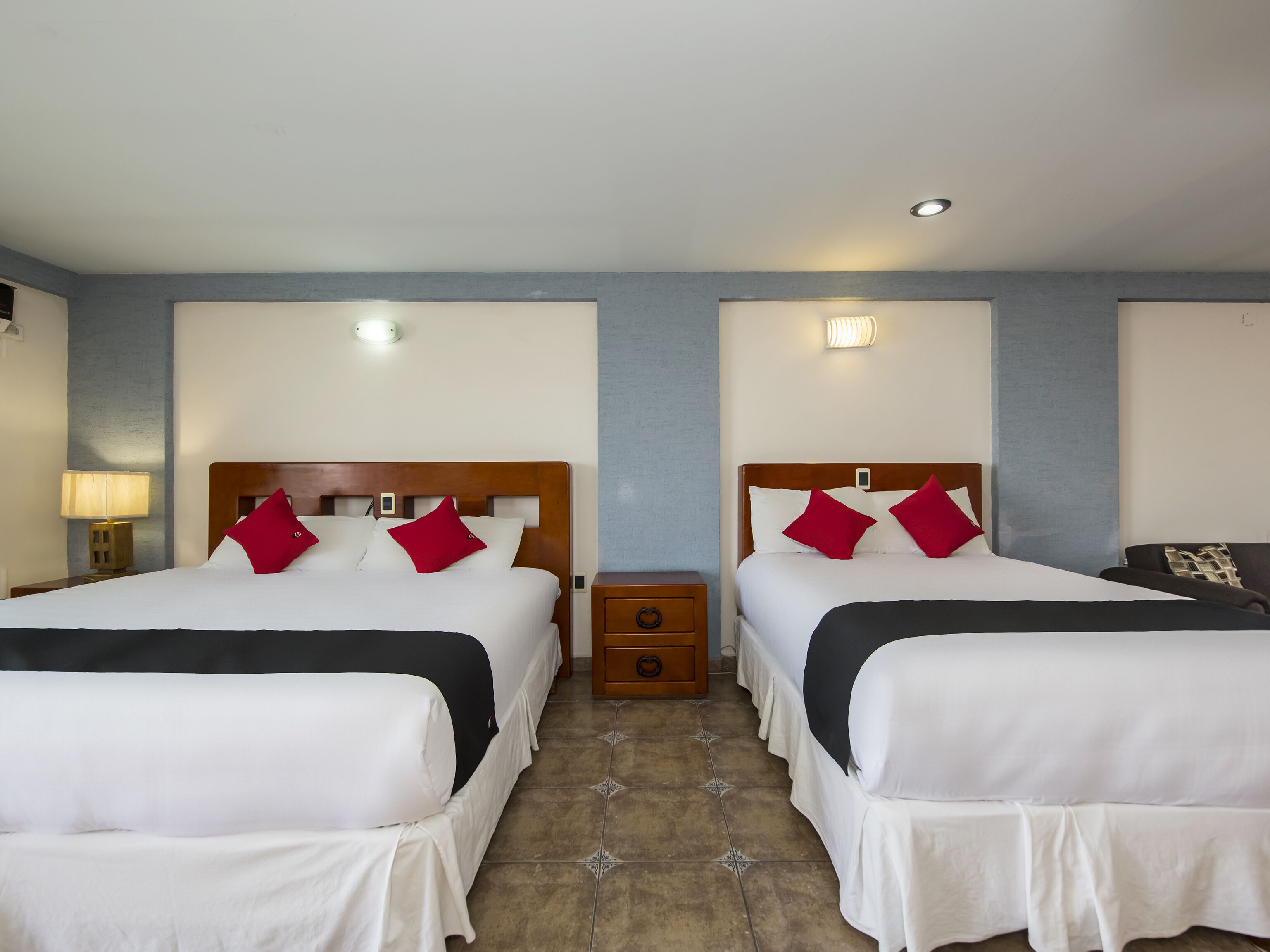 Une chambre d’hôtel avec deux lits, chacun doté de draps blancs et d’oreillers rouges, une table de chevet avec une lampe et un petit tiroir.
