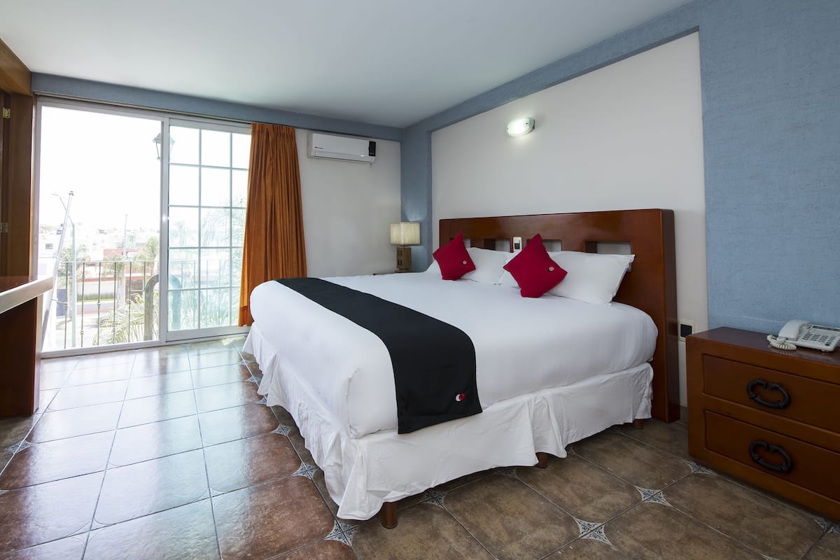 A hotel room with a large bed, a wooden nightstand, a telephone, and a view of the city through a large window.