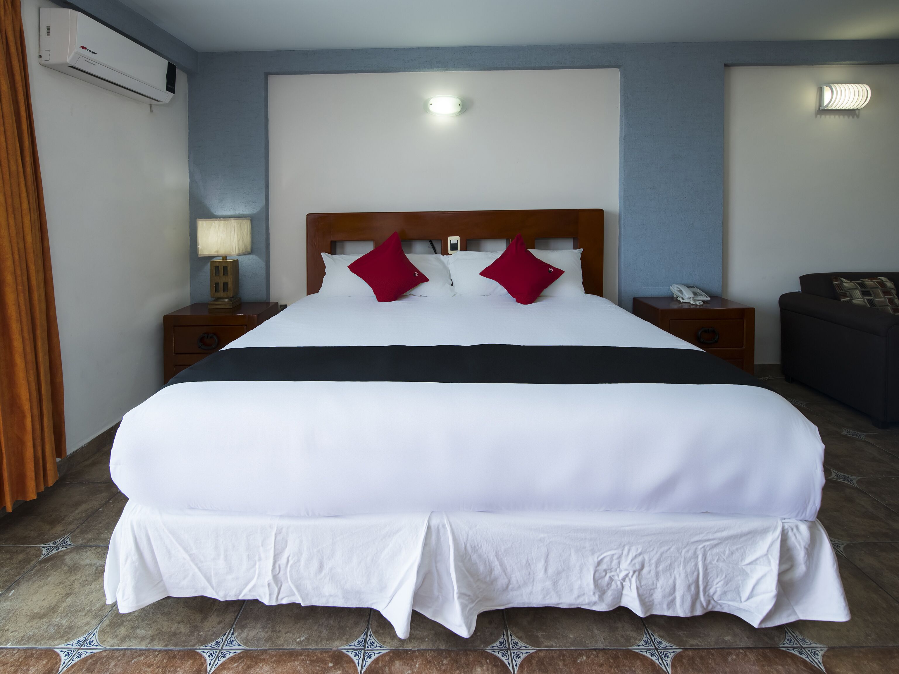 Habitación de hotel con una cama grande, dos mesitas de noche, una lámpara, un sofá y un aire acondicionado instalado en la pared.