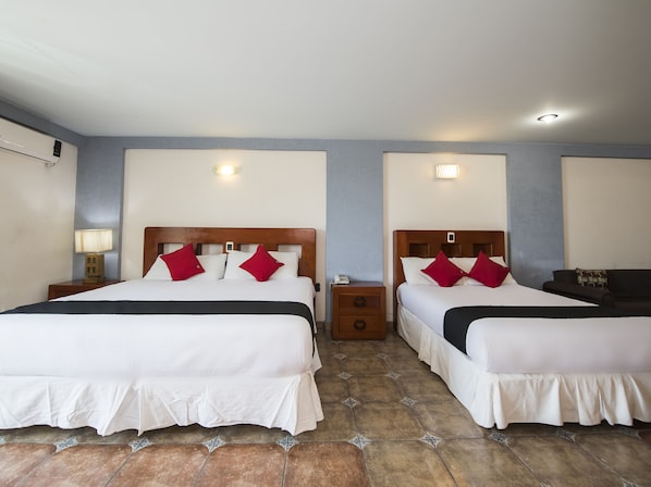 Habitación de hotel con dos camas, cada una con ropa blanca y almohadas rojas, una mesita de noche con lámpara y un cajón pequeño.