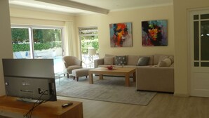 En moderne stue med et fladskærms-tv på et træstel, et beige sofasæt, et sofabord og et stort vindue med udsigt til en swimmingpool og grøn bevoksning.