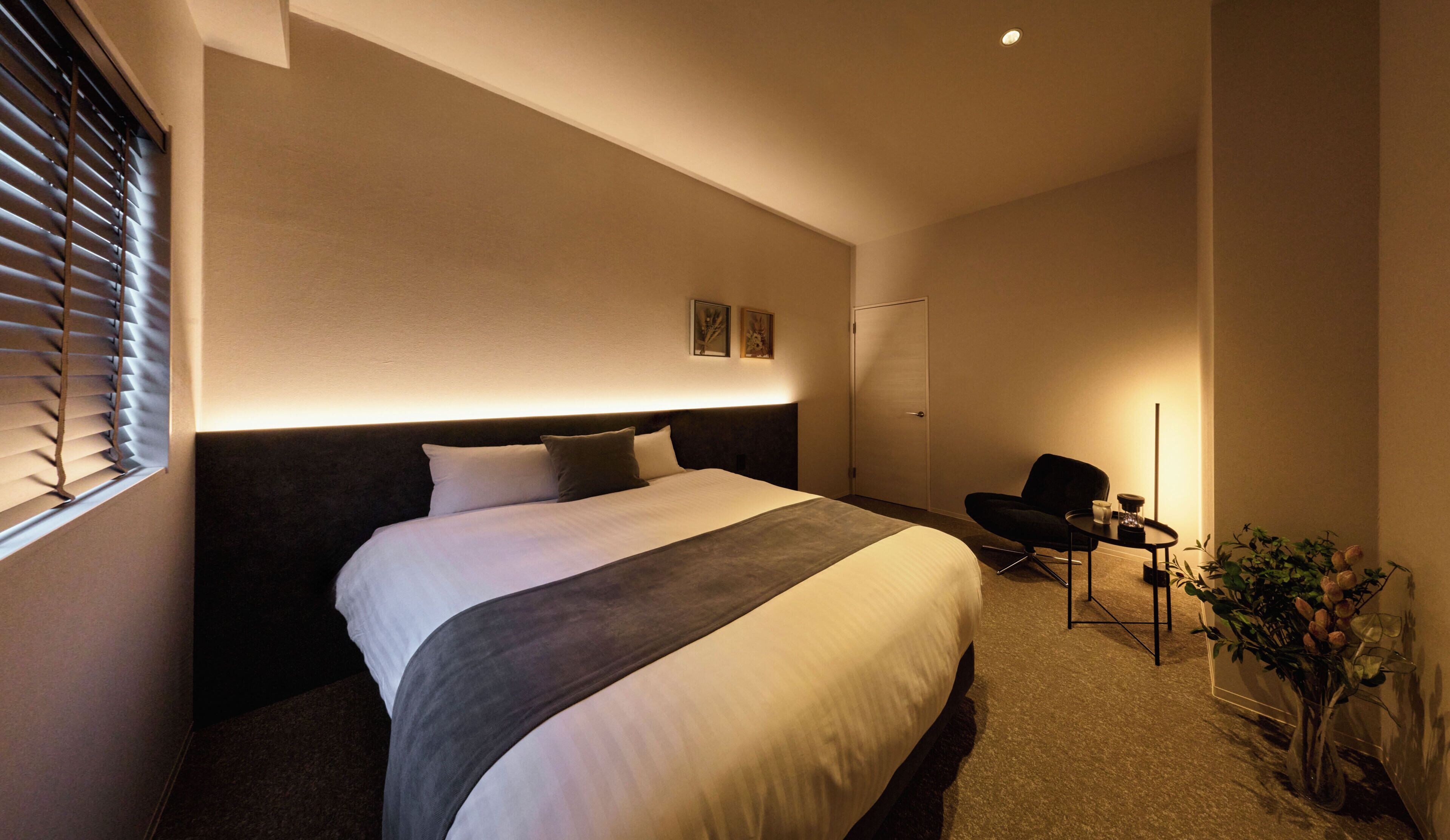 A hotel room with a bed, a chair, a small table, and a potted plant.