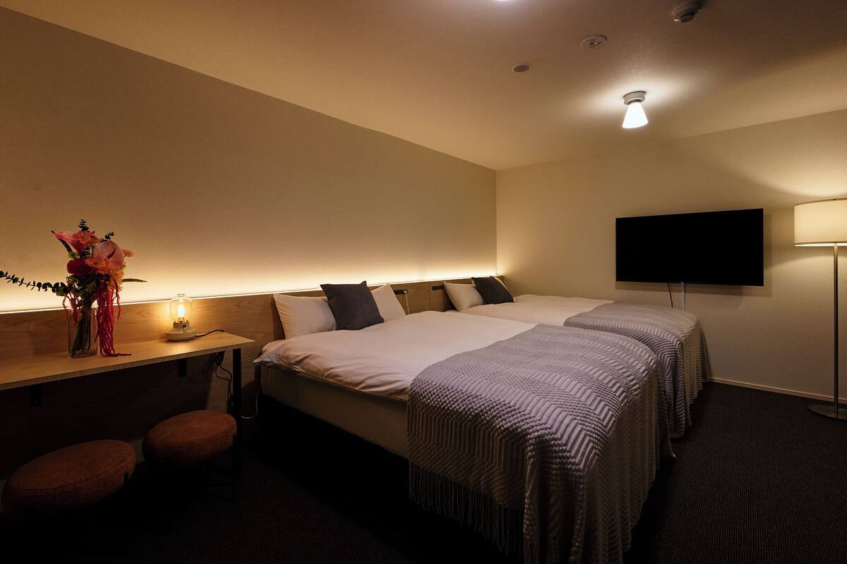 Habitación de hotel con dos camas, un escritorio con lámpara, televisión y un jarrón con flores.