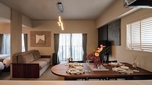 Une chambre d’hôtel moderne équipée d’un canapé, d’une table à manger pour quatre personnes, d’une télévision et d’une grande fenêtre avec des rideaux.