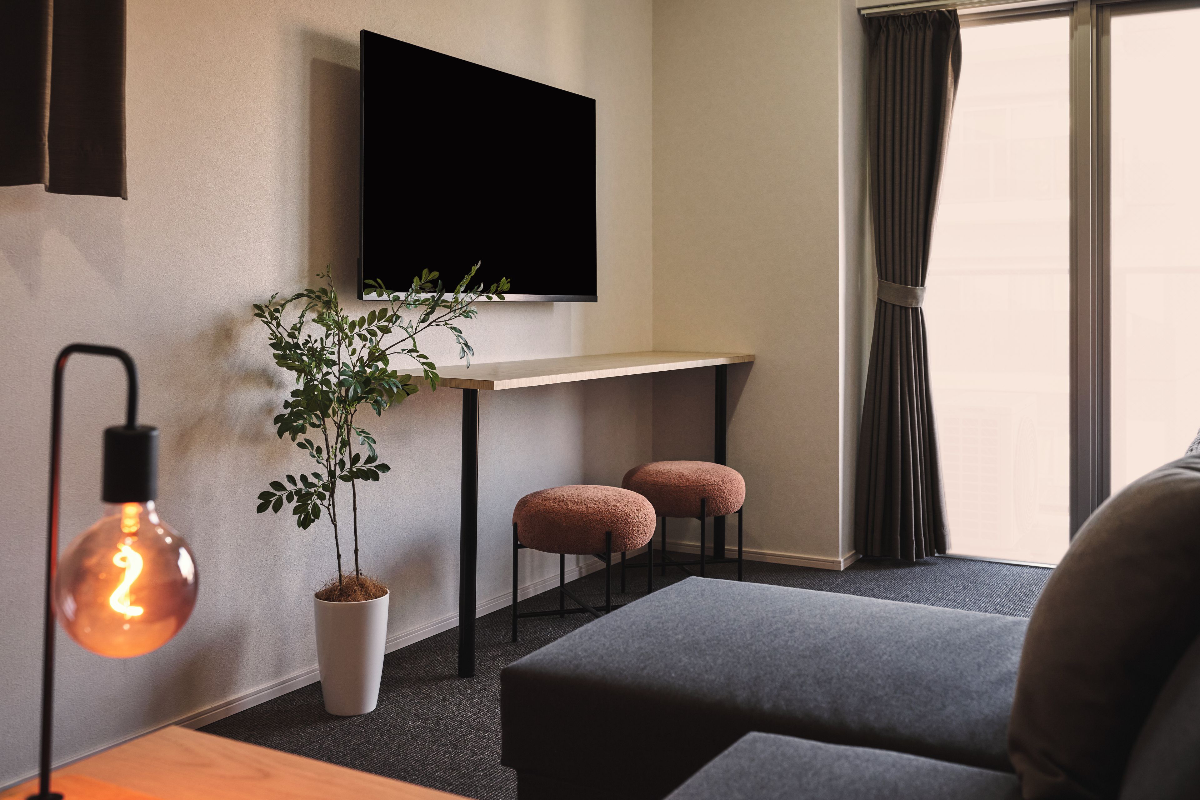 A modern living room with a flat-screen TV mounted on the wall, a potted plant, two cushioned stools, and a table with a lamp.