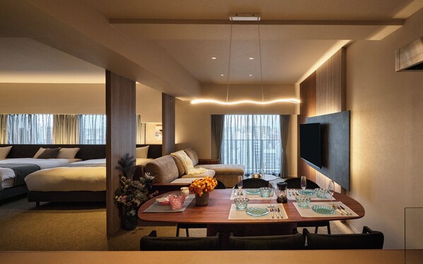 Une chambre d’hôtel comprenant un coin repas, un canapé, un téléviseur fixé au mur et une grande fenêtre avec des rideaux.