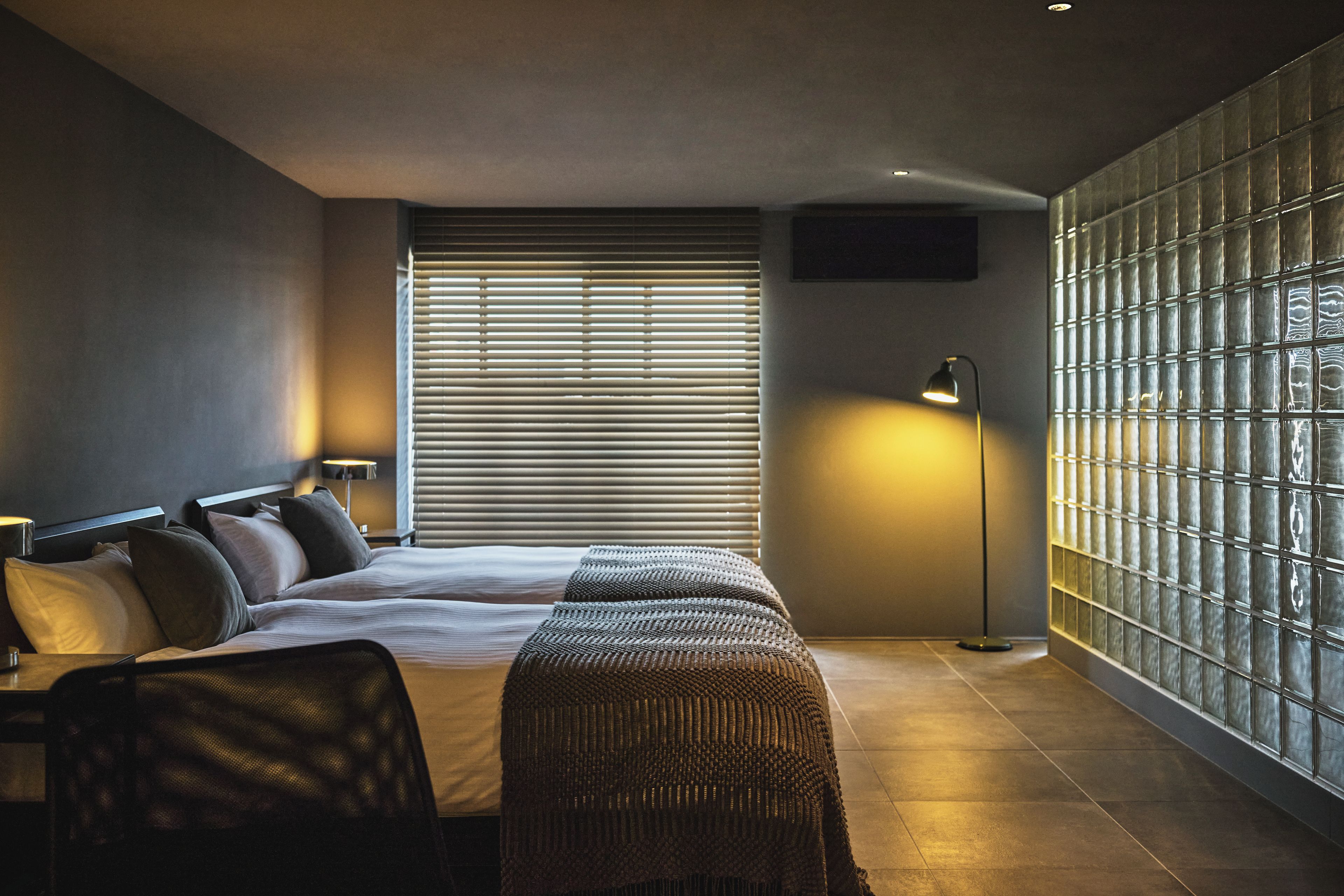 A modern hotel room with a large bed, two bedside tables, a standing lamp, and a view of the city through floor-to-ceiling windows.