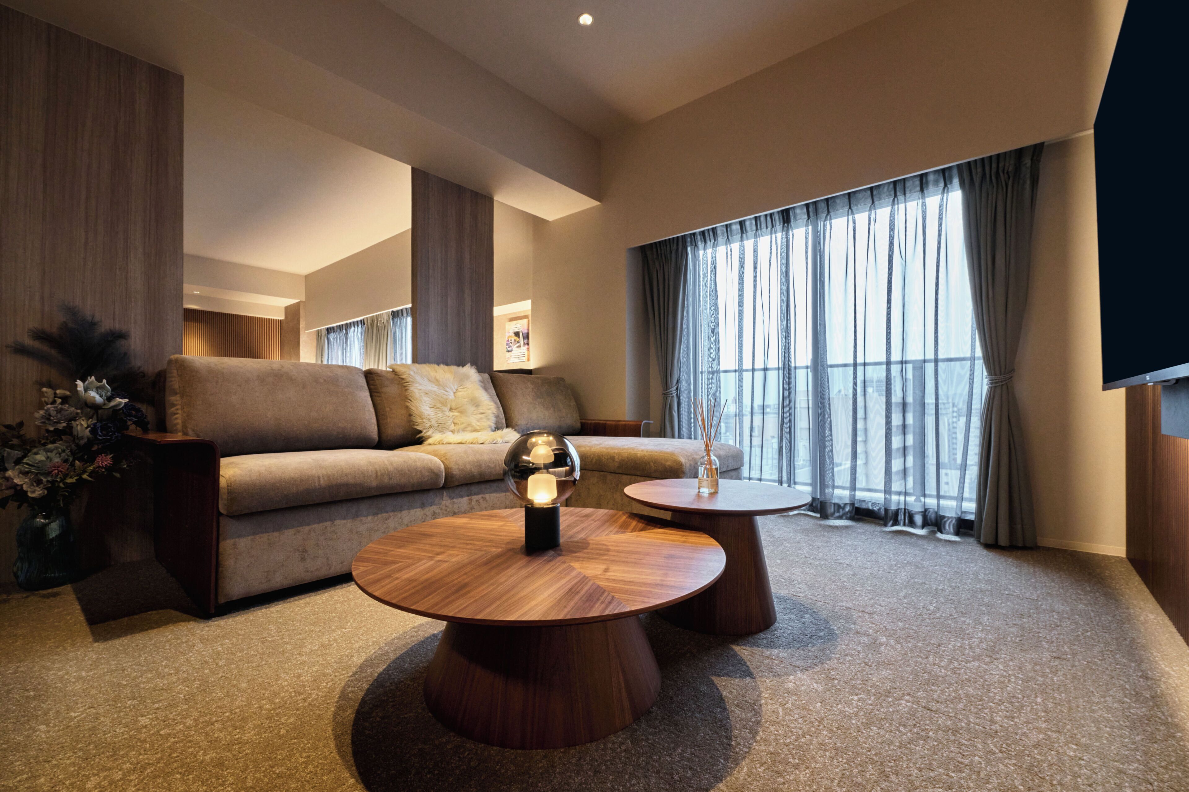 A modern living room with a sofa, two round wooden tables, a decorative vase, and a large window with curtains.