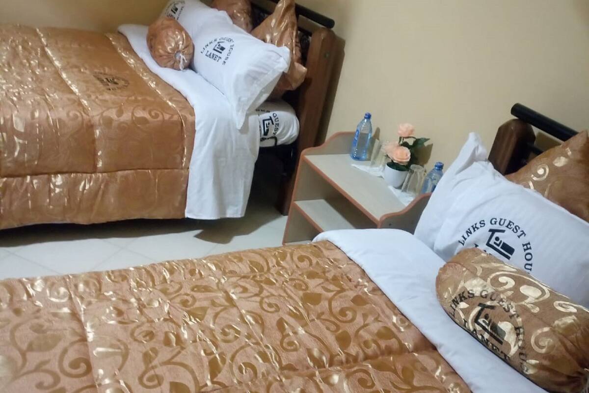 Una habitación de hotel con dos camas, cada una con ropa de cama estampada, una mesita de noche con una botella y una flor, y un buró con una lámpara.