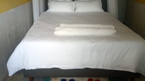 Een netjes opgemaakt bed met een witte spreideken en kussens, een paar blauwe en rode sloffen op een geruit vloerkleed, en een raam met witte gordijnen.