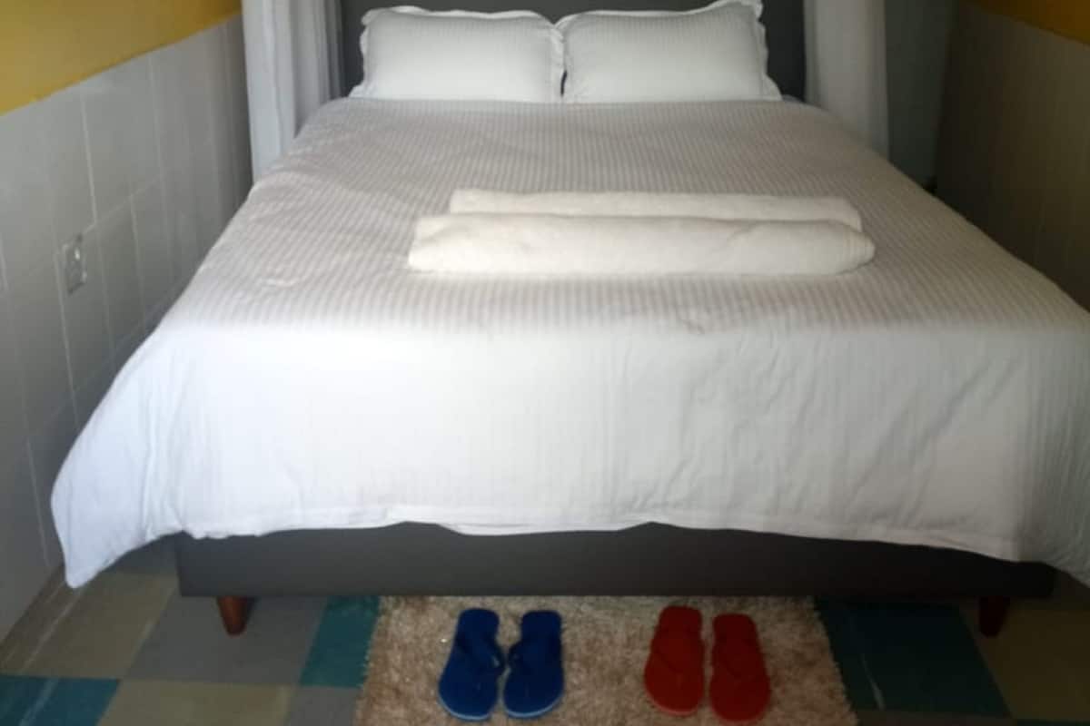 A neatly made bed with a white bedspread and pillows, a pair of blue and red slippers on a checkered rug, and a window with white curtains.