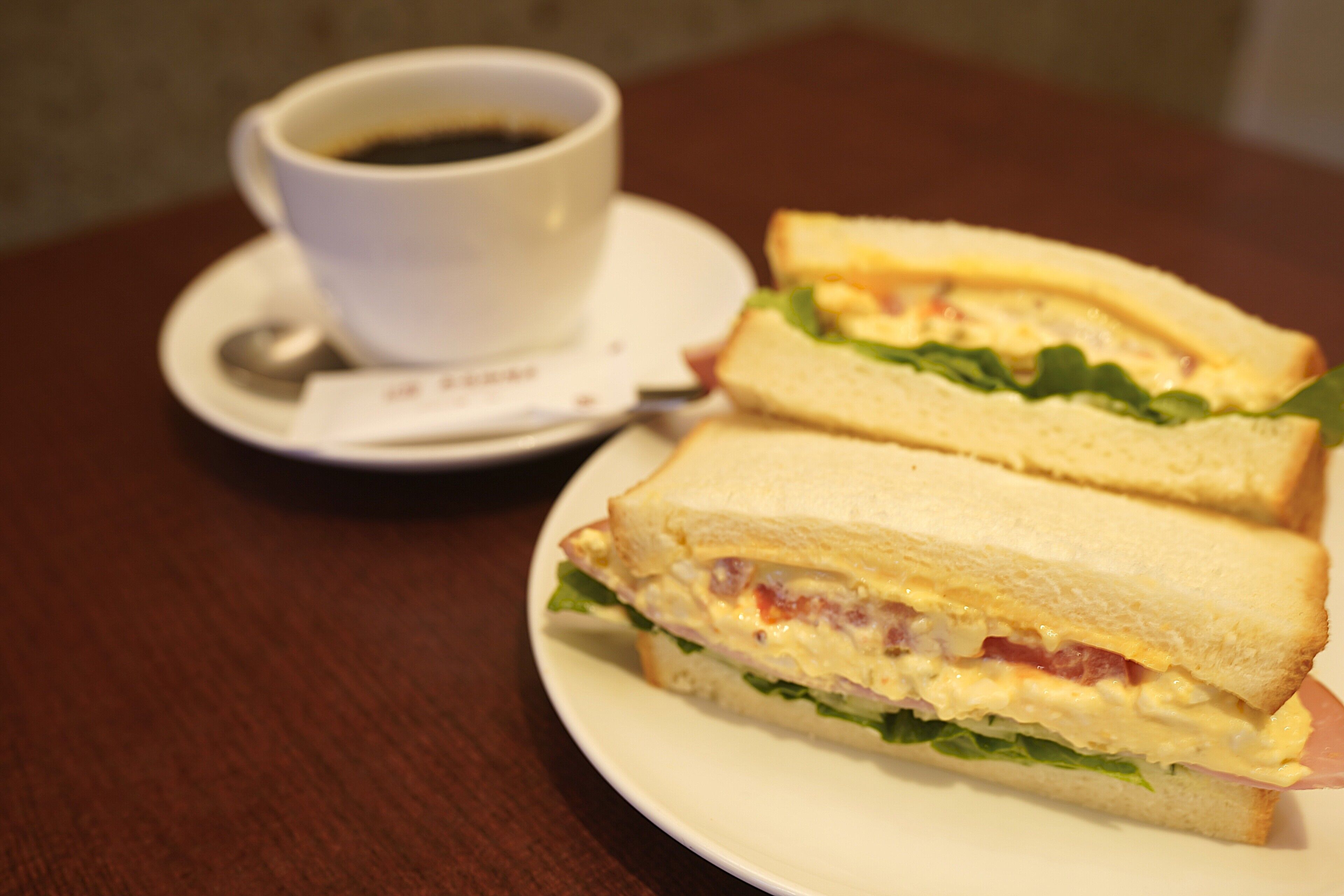 A sandwich on a plate with a cup of coffee in the background.