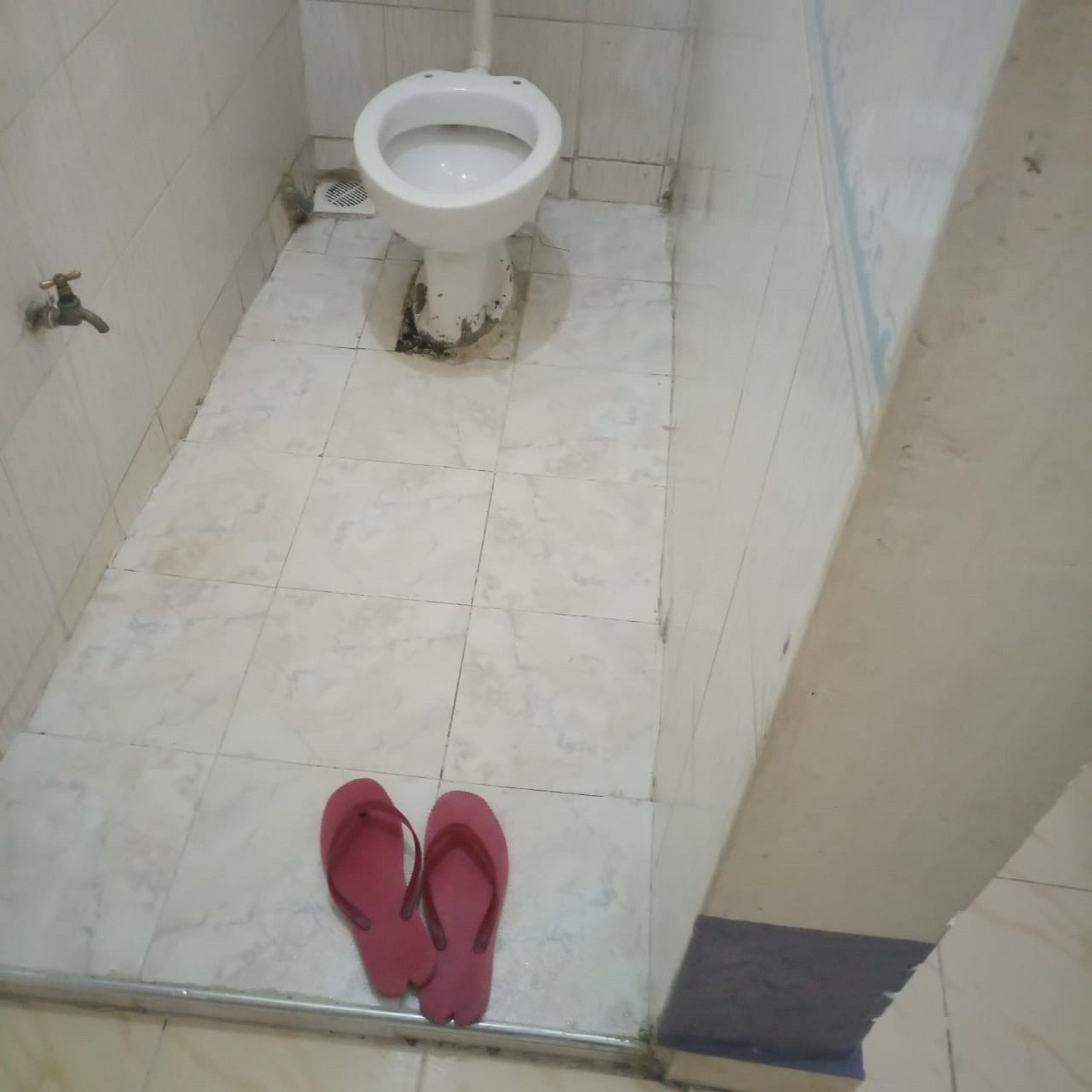 A bathroom with a toilet, a drain, and a pair of red slippers on the floor.