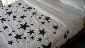 A bed with a white and black star-patterned bedspread, a wooden nightstand with a lamp, and sheer curtains. - Hudson's Sports Bar and Restaurant (Nakuru)