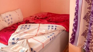 Une petite chambre avec un lit simple, un mur uni, une couvre-lit à motifs et un rideau orné d’un motif géométrique.