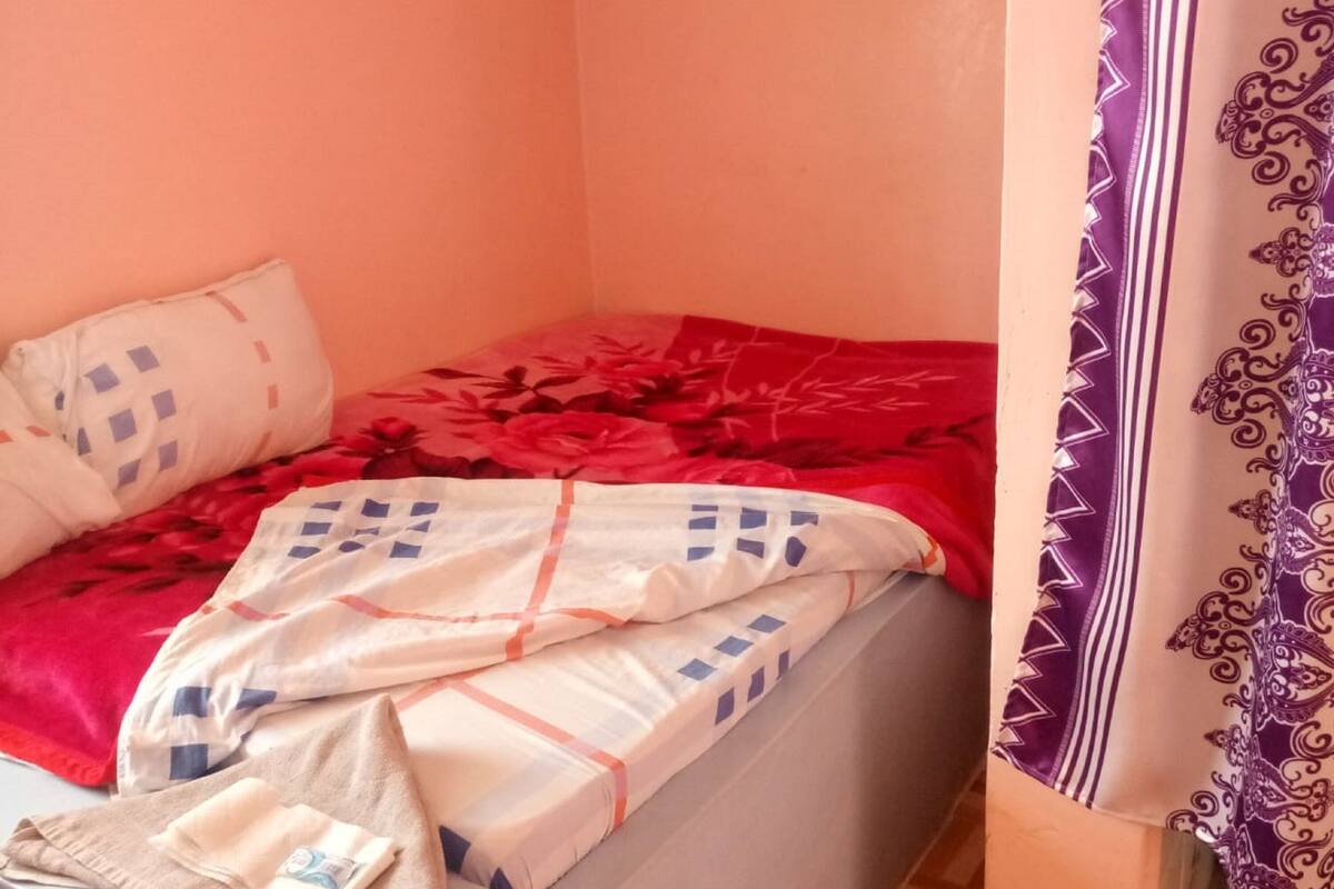 A small, single-bed room with a plain wall, a patterned bedspread, and a curtain with a geometric design.