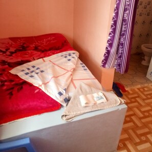 A bed with a red blanket and a beige pillow with a blue pattern, a small towel, and a remote control on a wooden floor. - Duplex Lodge (Nakuru)