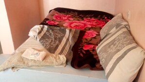 Un lit avec plusieurs oreillers et une couverture, une chaise en plastique bleue et un sol carrelé.