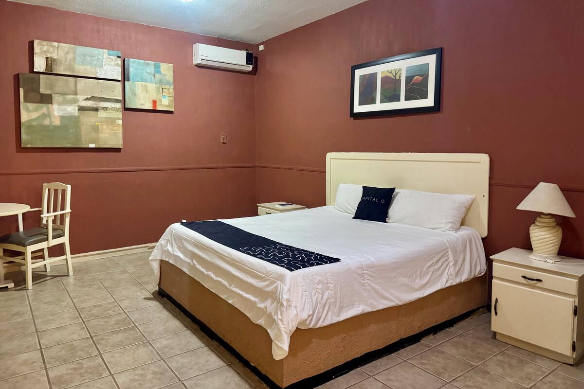 A hotel room with a bed, a chair, a bedside table, a lamp, and artwork on the walls.