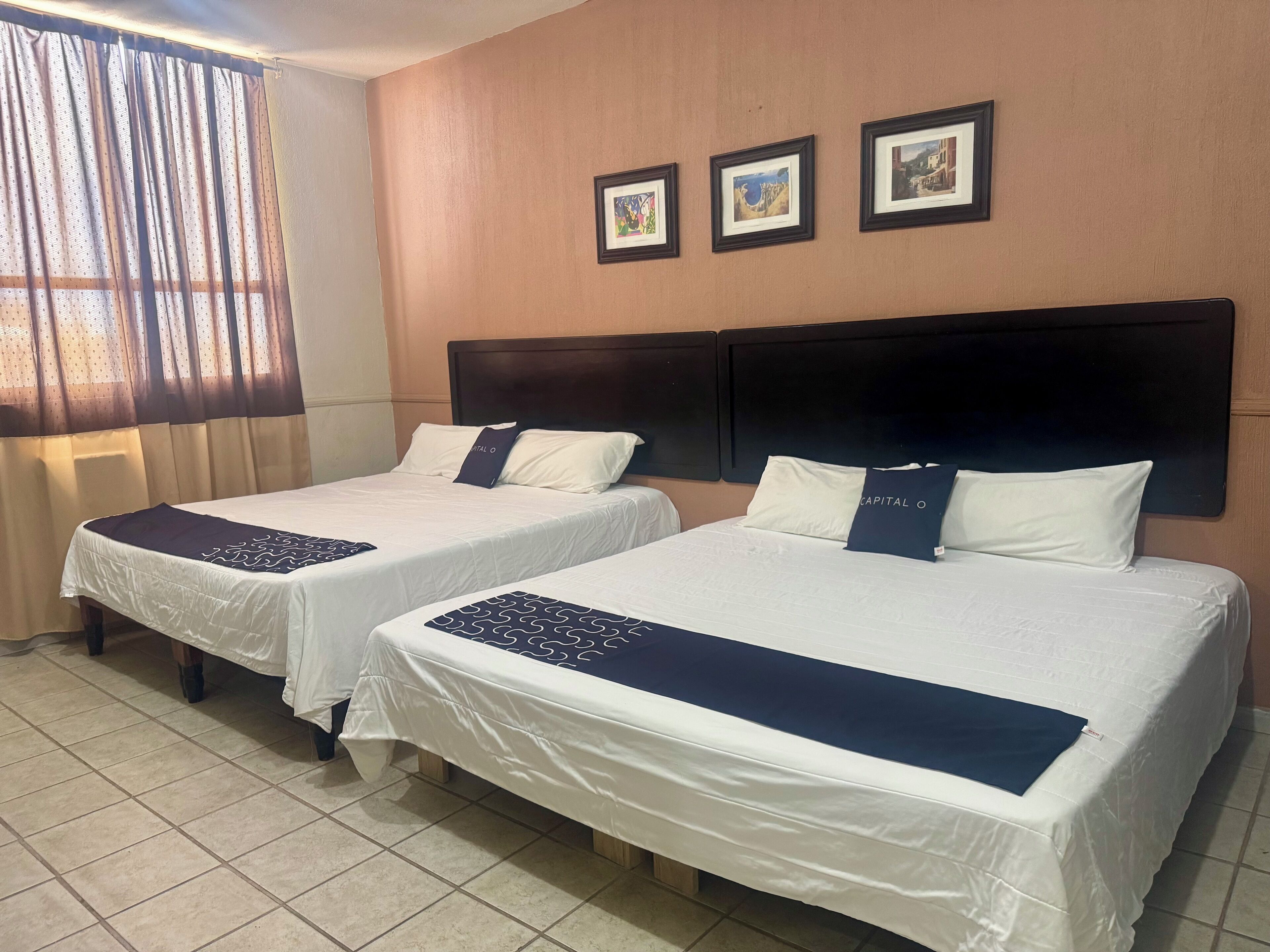 A hotel room with two beds, a large window with curtains, and three framed pictures on the wall.
