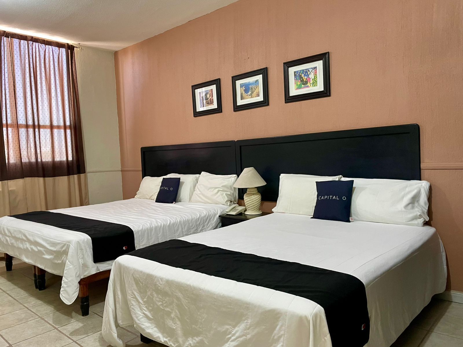 Una habitación de hotel con dos camas, una mesita de noche con lámpara y tres cuadros enmarcados en la pared.