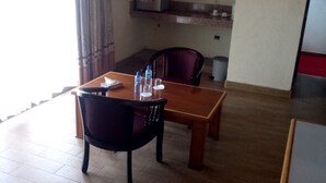 A hotel room with a wooden dining table, two chairs, a small shelf with a box, and a trash bin. - Top Cliff (Nakuru)