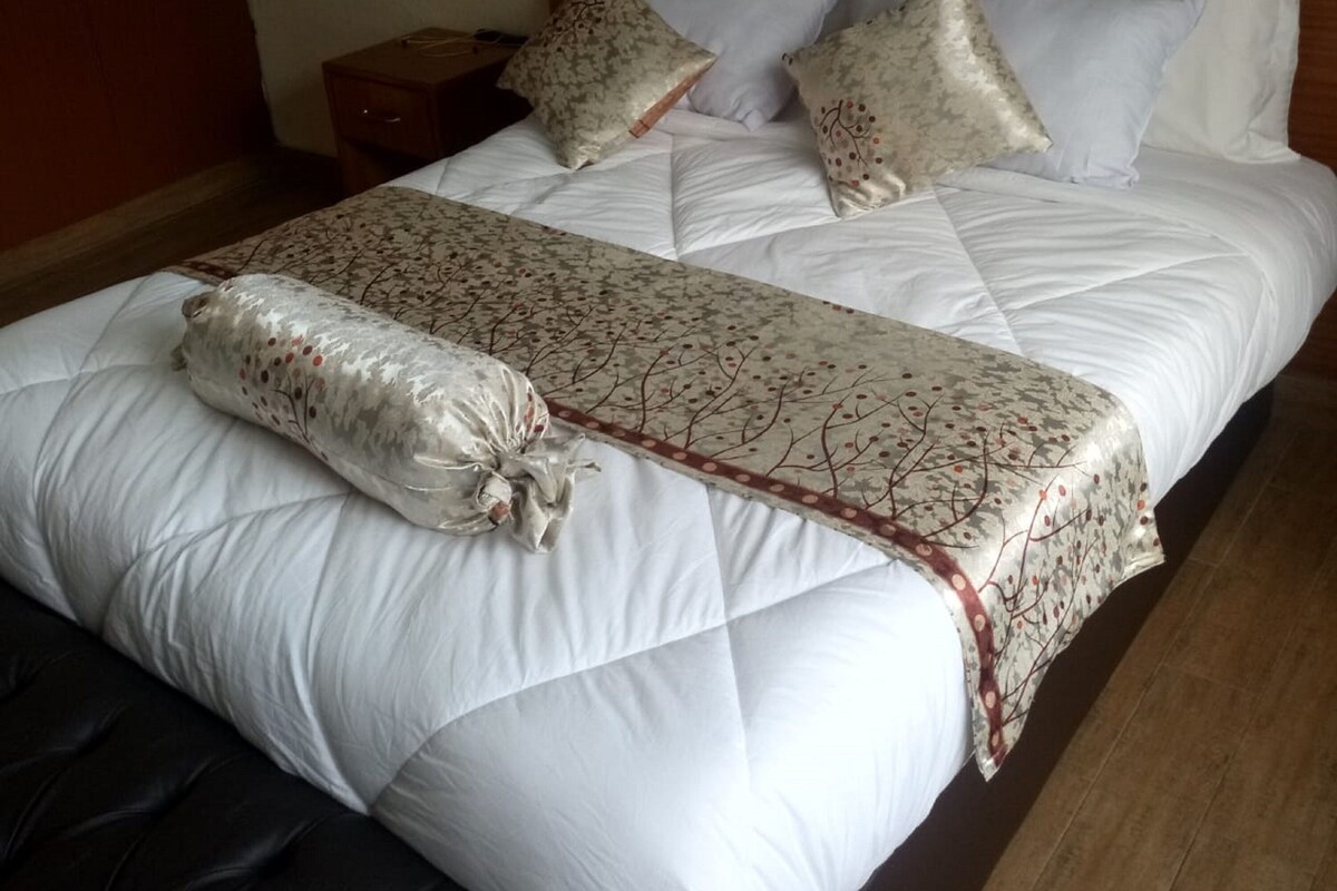 Cama bem arrumada com manta acolchoada, travesseiro decorativo e mesa de cabeceira com abajur.