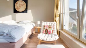 A hotel room with a bed, a chair with a colorful cushion, a bedside table with a lamp, and a large window with curtains. - Luxury Pool Villa in Sosoye (Anhée)