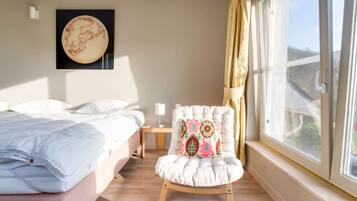 A hotel room with a bed, a chair with a colorful cushion, a bedside table with a lamp, and a large window with curtains.